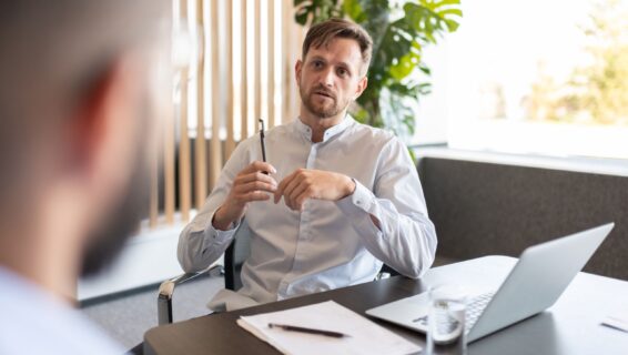 Ein Mann sitzt am Konferenztisch mit dem Laptop und Block vor sich und ist im Gespräch mit einer zweiten Person die man von hinten sieht.