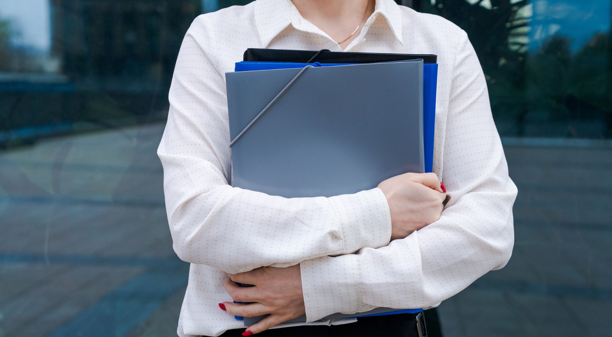 Eine Frau hält mehrere Bewerbungsmappen im Arm und steht vor einem Bürogebäude.