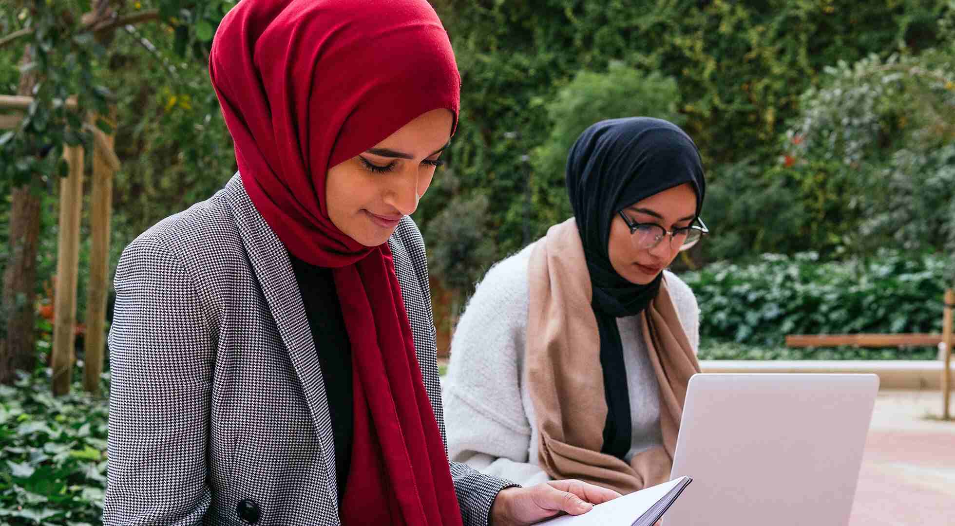 Zwei junge Frauen mit Kopftuch sitzen in einem Garten und arbeiten konzentriert an ihren Laptops.