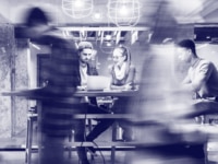 odernes Büro mit Mitarbeitern: Zwei Personen arbeiten gemeinsam an einem Laptop, während andere Kollegen im Hintergrund in Bewegung sind. Schwarzweiß-Fotografie mit dynamischer Bewegungsunschärfe.