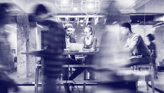 odernes Büro mit Mitarbeitern: Zwei Personen arbeiten gemeinsam an einem Laptop, während andere Kollegen im Hintergrund in Bewegung sind. Schwarzweiß-Fotografie mit dynamischer Bewegungsunschärfe.